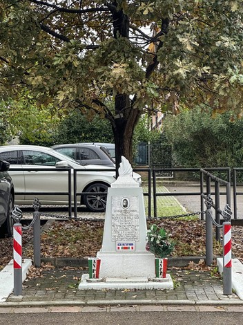 Cippo commemorativo dedicato a Bellotti, circondato da piante e veicoli, in un'area verde.