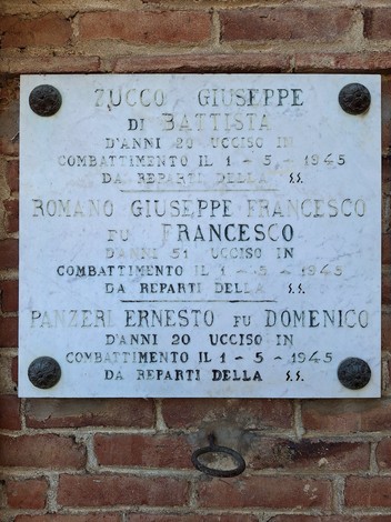 Lapide marmorea con iscrizione, fissata a una parete in mattoni con quattro rosette angolari.