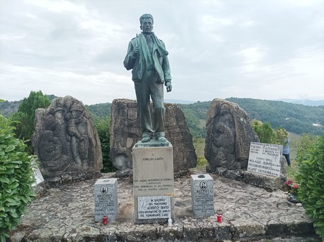 Monumento ai caduti con statua e pietre commemorative, immerso nel verde e panoramica su un paesaggio naturale.