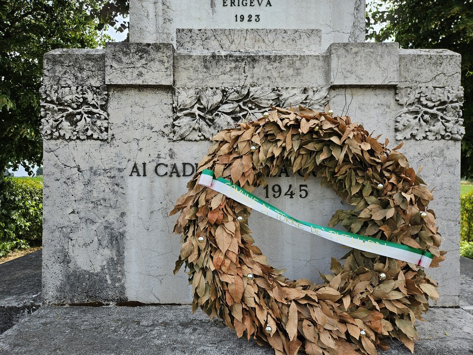 Monumento ai Caduti con corona di alloro secca e decorazioni floreali, simbolo di commemorazione.
