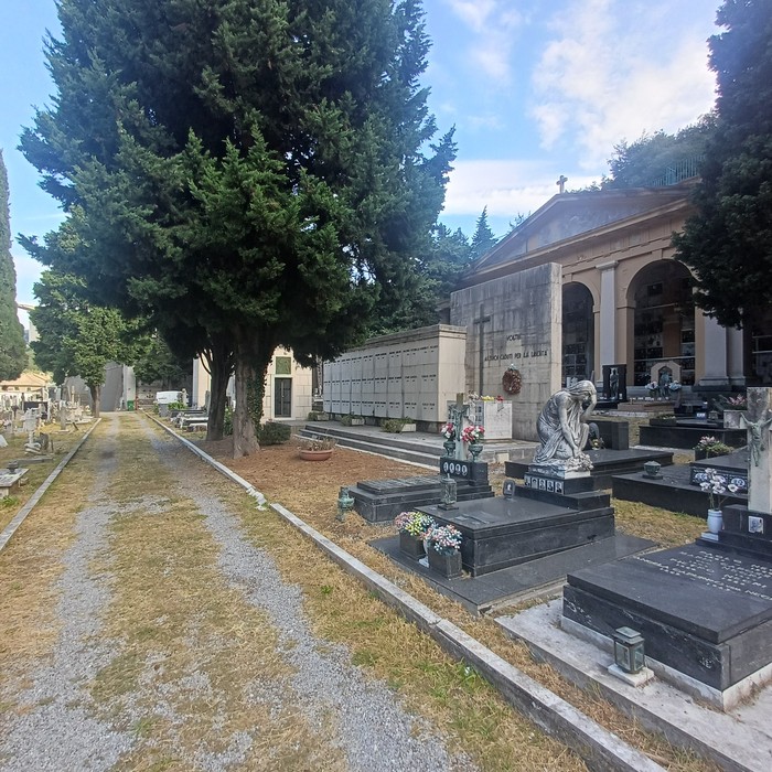 Viale di cimitero con cappelle e tombe in marmo nero, statua di donna china e cipressi sullo sfondo.