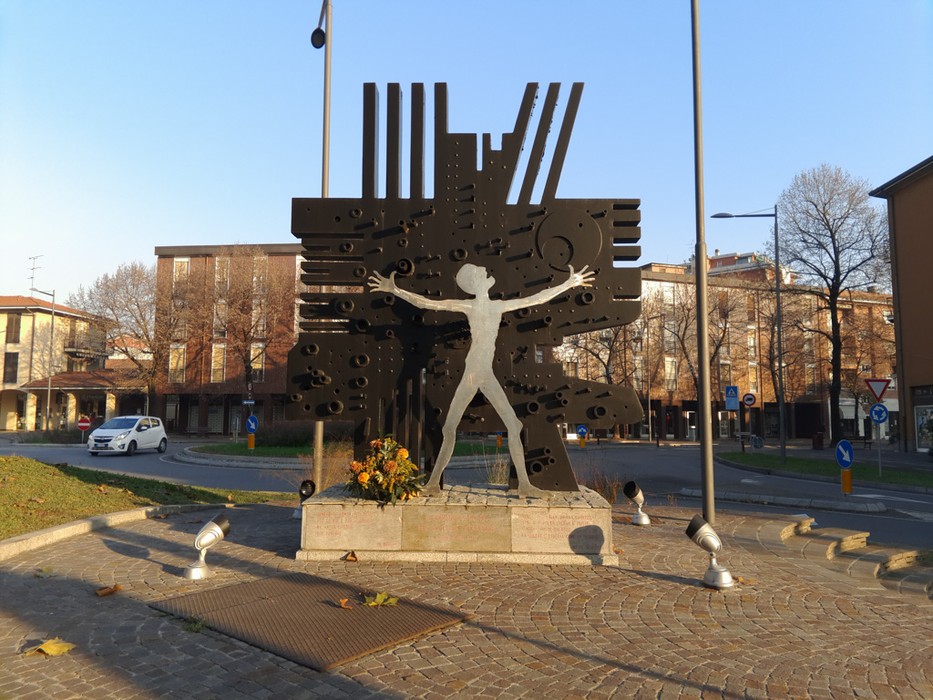 Monumento in metallo scuro raffigurante una figura umana stilizzata a braccia aperte, su rotonda cittadina.