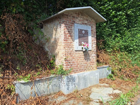 Monumento a Zulian, struttura in mattoni con plaquette commemorativa e fiori, circondata da vegetazione.