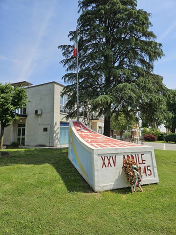 Monumento al XXV Aprile, con bandiera e decorazioni floreali, immerso nel verde urbano.