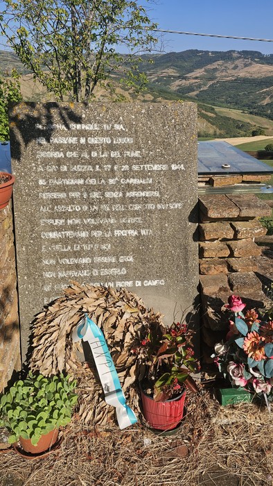 Monumento dedicato a Ca' di Guzzo, circondato da piante e fiori in un paesaggio collinare.