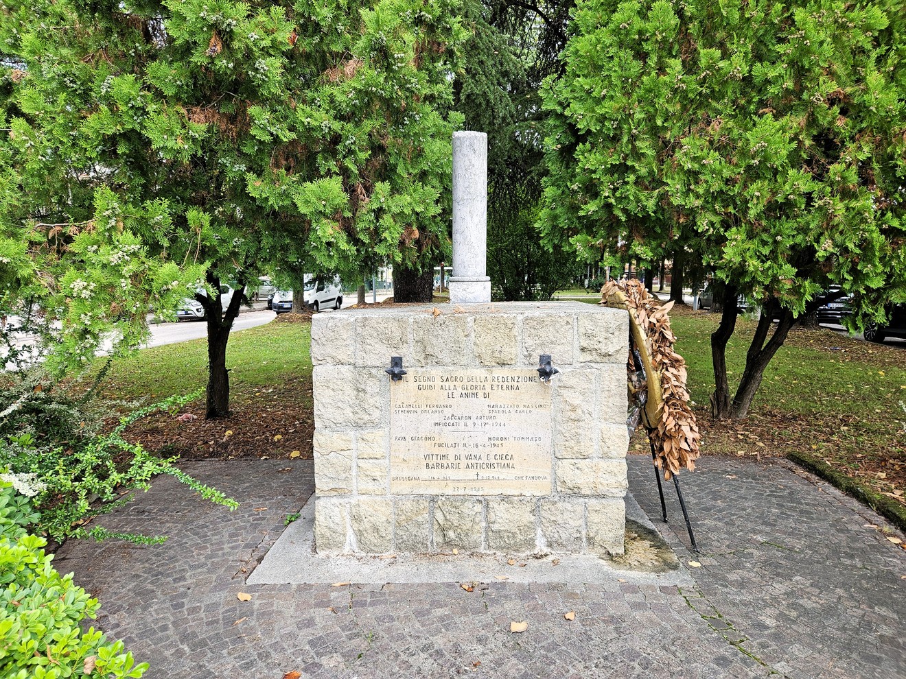 Cippo in pietra con colonna e iscrizione, collocato in un'aiuola alberata di un parco.
