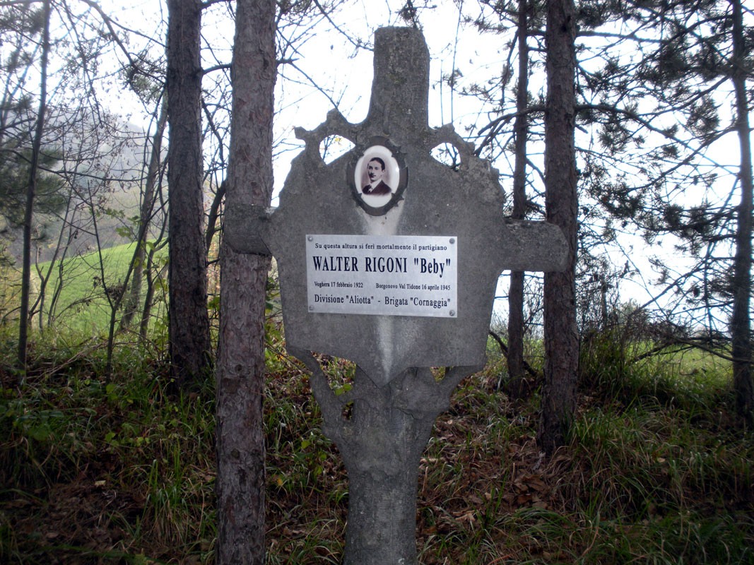 Targa commemorativa di Walter Rigoni, situata in un bosco, con una foto storica e informazioni biografiche.