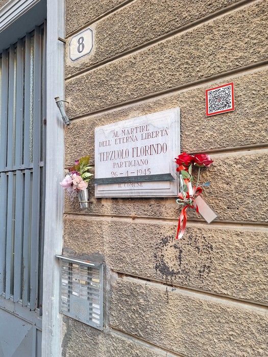 Lapide in marmo su parete esterna, con iscrizione e fiori freschi deposti sotto, accanto al civico 8.