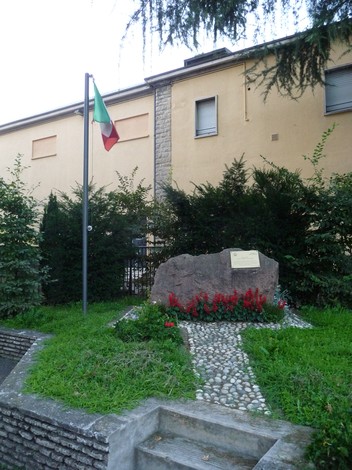 Monumento alla Resistenza con bandiera italiana, circondato da alberi e fiori, in un contesto urbano tranquillo.