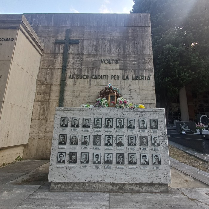 Parete in pietra con croce e iscrizione, lapide con foto e nomi dei caduti, fiori davanti.