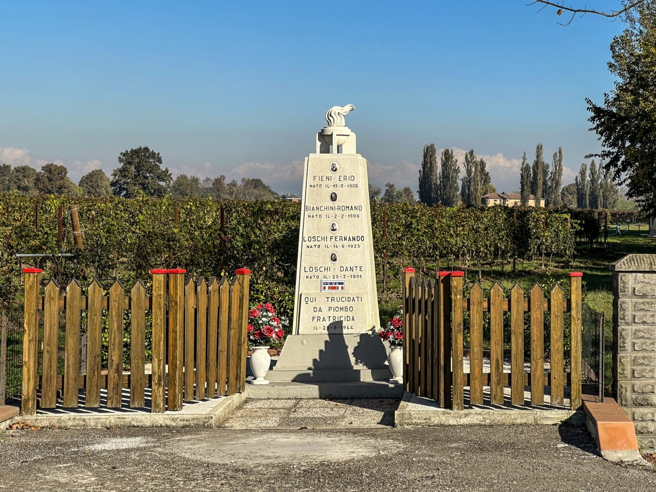 Monumento ai Loschi, Fieni e Bianchini, situato in un'area verde con un recinto di legno e decorazioni floreali.