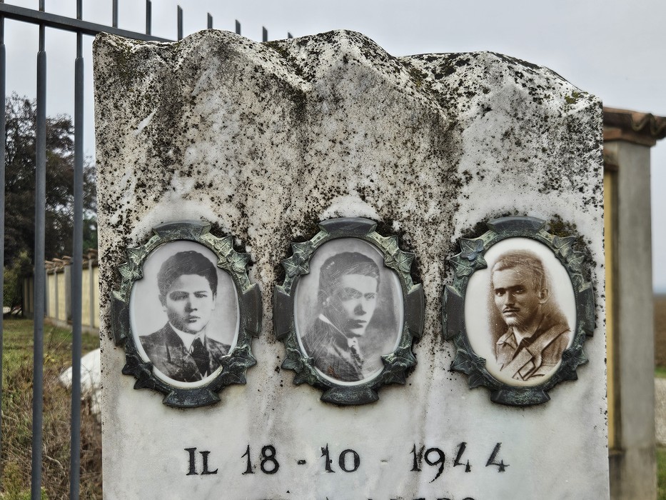 Cippo in pietra con tre ritratti ovali di giovani uomini e iscrizione, dietro una recinzione metallica.