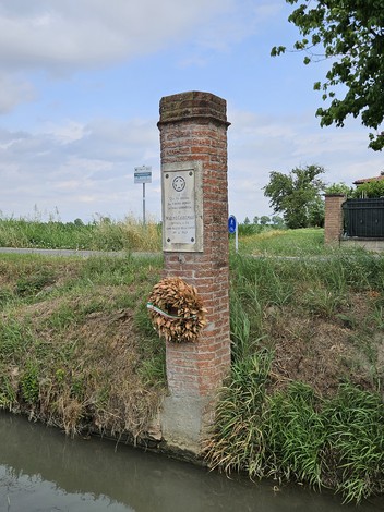 Cippo in pietra con corona di fiori, immerso nella natura vicino a un canale e una strada.