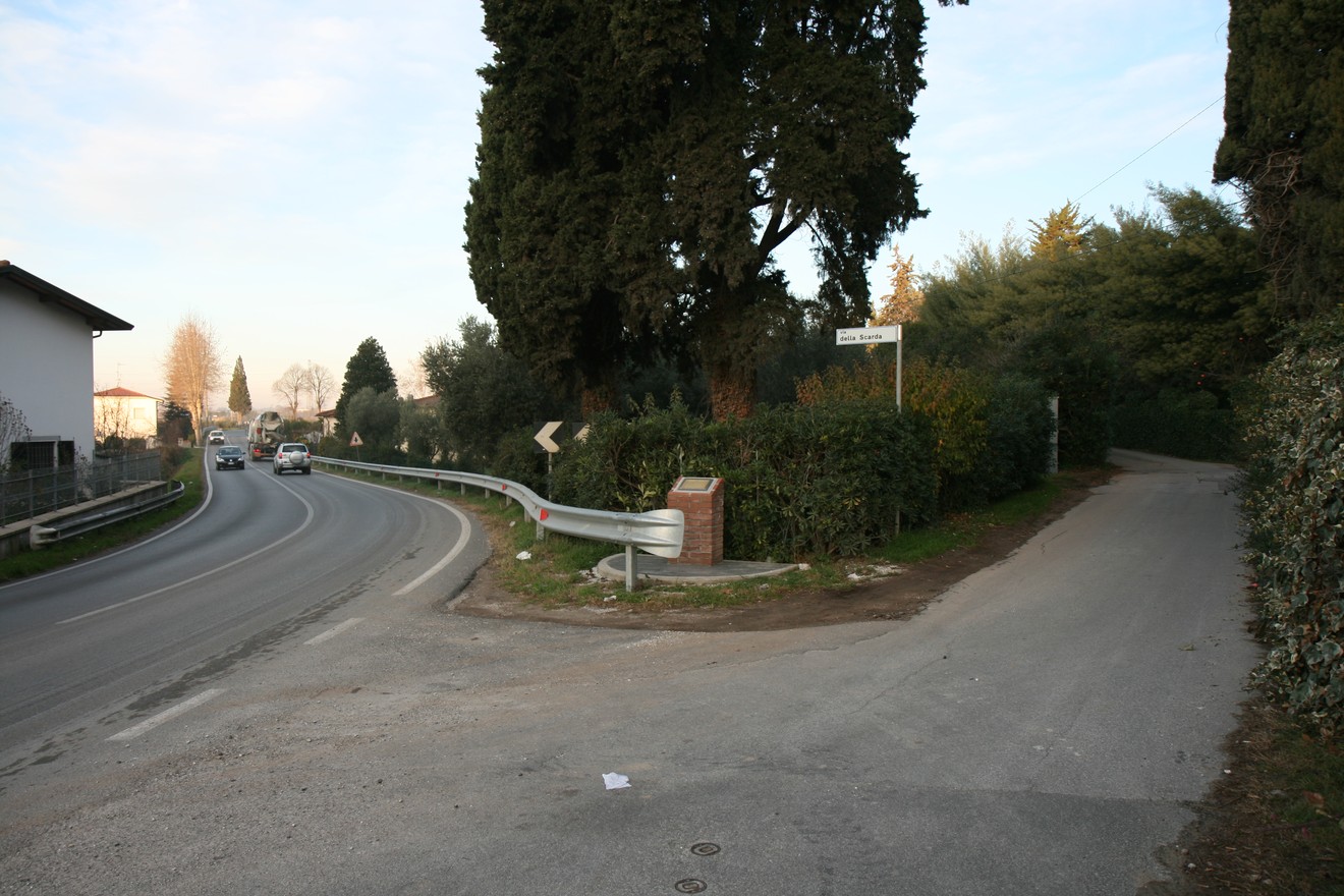 Cippo di Sassaia all'incrocio tra strada principale e viale alberato, circondato da vegetazione.