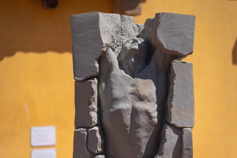 Statua in pietra grigia con figura scavata al centro, su sfondo di parete gialla.