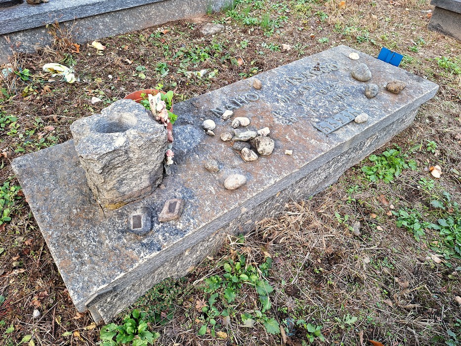 Tomba in pietra con iscrizione, sassi deposti sopra e una piccola cavità scavata su un lato.