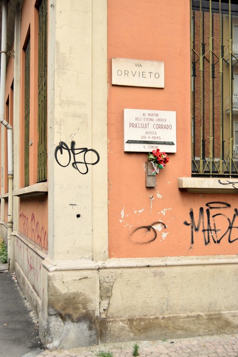Lapide commemorativa a Prassuit, angolo di strada con fiori in omaggio e scritte sui muri.