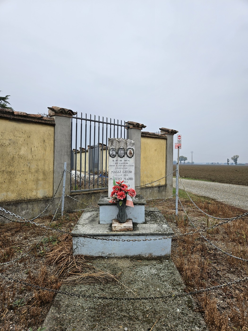Cippo in pietra con iscrizione e fotografie, fiori ai piedi, lungo un muro di recinzione su strada sterrata.