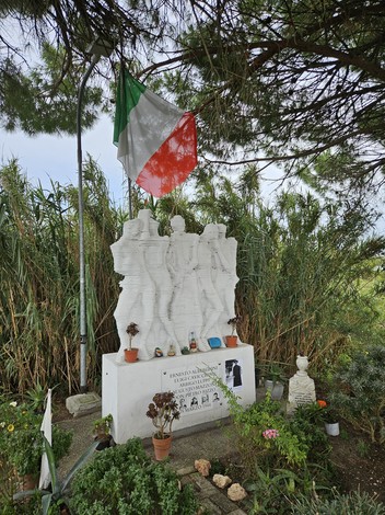 Monumento ai Martiri della Macchinina, raffigurante quattro figure in marmo tra piante e fiori. Bandiera italiana presente.