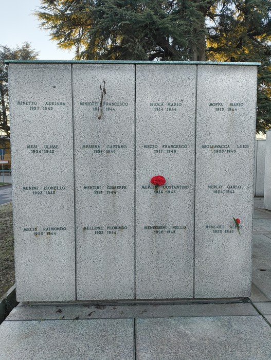 Monumento commemorativo con nomi incisi, fiori rossi e alberi sullo sfondo, simbolo di memoria e rispetto.