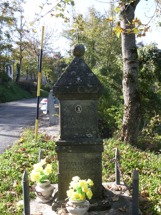 Cippo commemorativo in pietra, circondato da fiori, situato lungo una strada immersa nella natura.