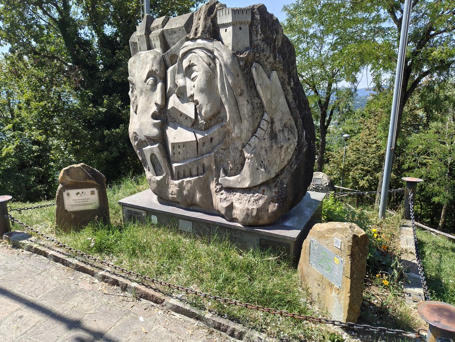 Scultura in pietra con volti e scene in rilievo, su basamento, circondata da alberi e due lapidi laterali.
