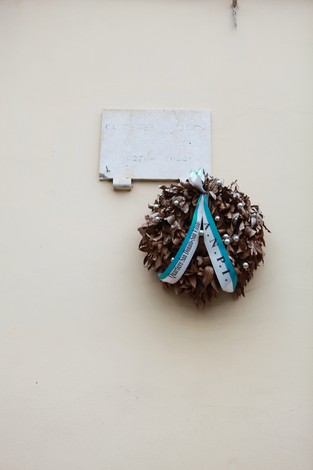 Lapide commemorativa con corona di alloro, appesa a un muro chiaro in un contesto urbano.