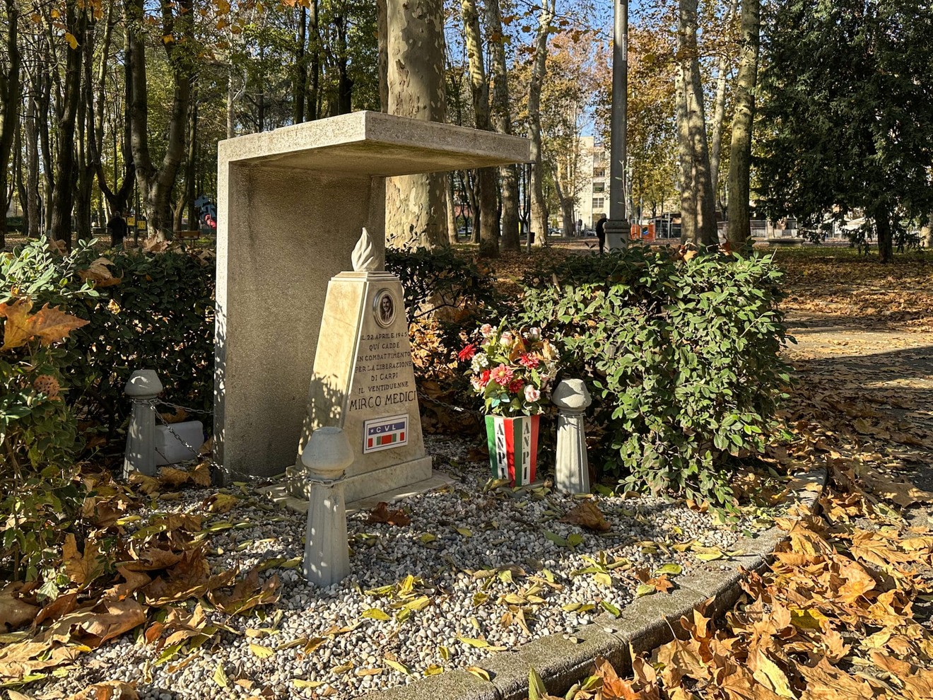Cippo commemorativo dedicato ai Medici, circondato da piante e fiori, in un parco durante l'autunno.