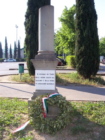 Cippo commemorativo dedicato ai caduti, adornato da una corona di alloro e circondato da alberi.