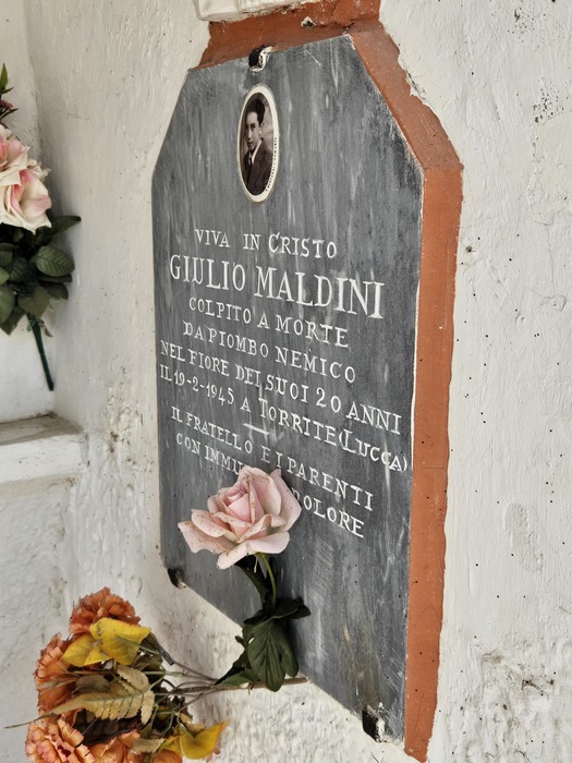 Lapide in marmo scuro con foto e iscrizione, fissata a un muro, con fiori ai piedi.
