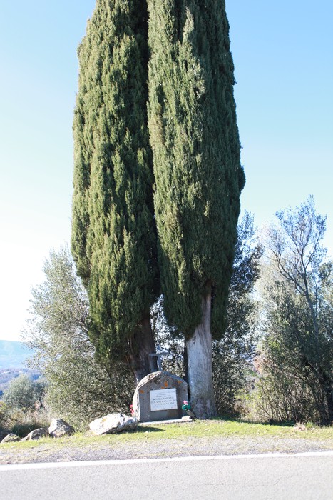 Cippo commemorativo circondato da cipressi in un contesto naturale, lungo una strada panoramica.