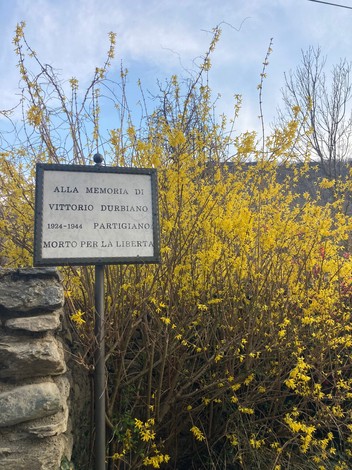 Lapide commemorativa a Durbiano circondata da fiori gialli, in memoria di un partigiano.