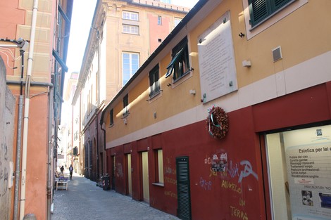 Lapide commemorativa su un muro di un vicolo, con edifici colorati e scritte sui muri circostanti.
