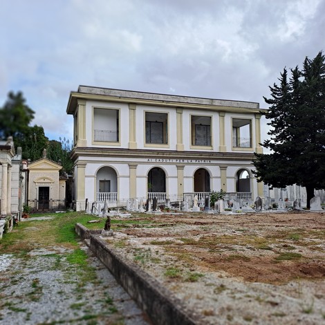 Edificio a due piani con portico ad arcate e iscrizione, circondato da lapidi in un cimitero.