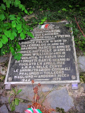 Lapide commemorativa dedicata ai Lojodice e altri, situata in un contesto naturale, con decorazioni floreali.