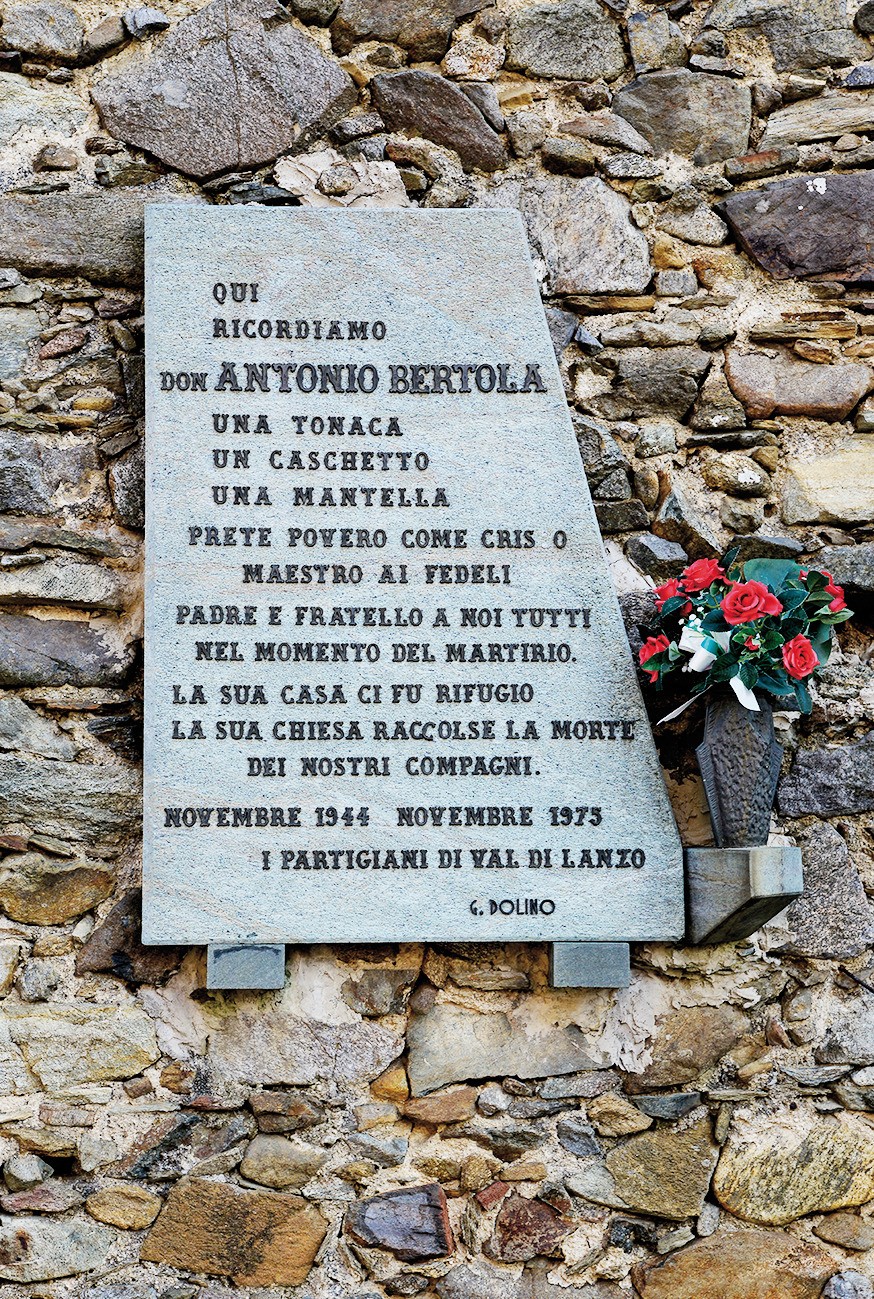 Lapide in marmo con iscrizione su muro in pietra, con mazzo di rose rosse e croce accanto.
