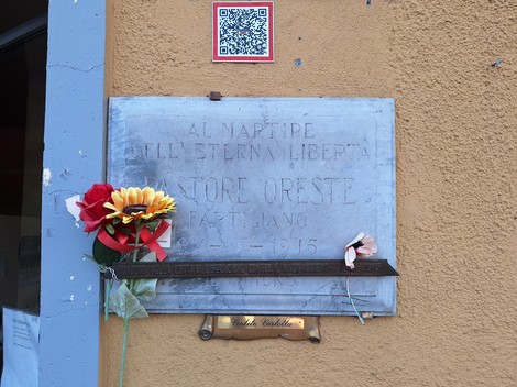 Lapide in marmo su parete esterna, con iscrizione e fiori finti deposti sotto.