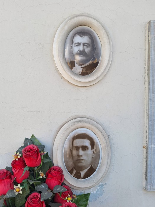 Lapide commemorativa con due ritratti fotografici e un mazzo di rose rosse.