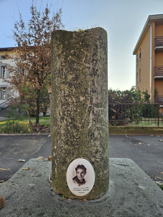Cippo commemorativo con effigie e iscrizioni, circondato da un contesto urbano e vegetazione.