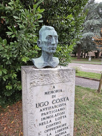 Busto in bronzo di Ugo Costa su un piedistallo commemorativo, circondato da vegetazione.