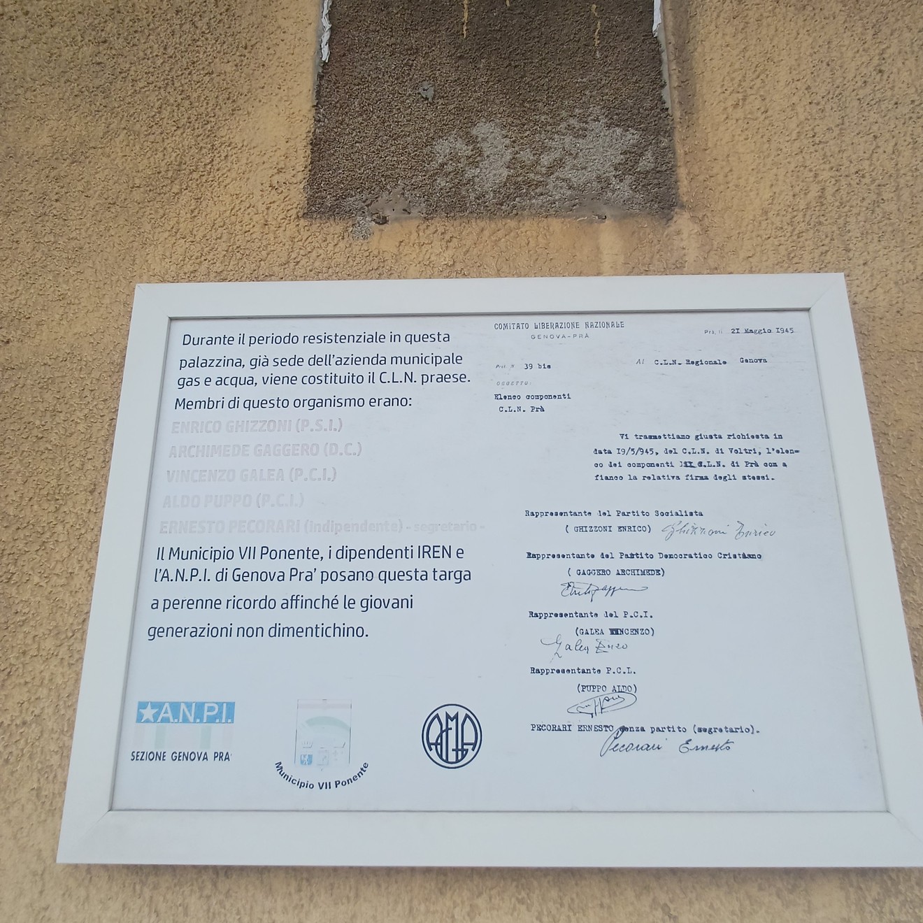 Targa bianca con iscrizione, affissa su una parete esterna accanto a un documento incorniciato.