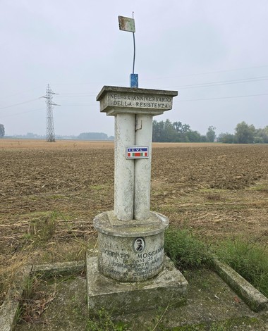 Cippo commemorativo a Moscardini, con bandiera e contesto rurale alle spalle.