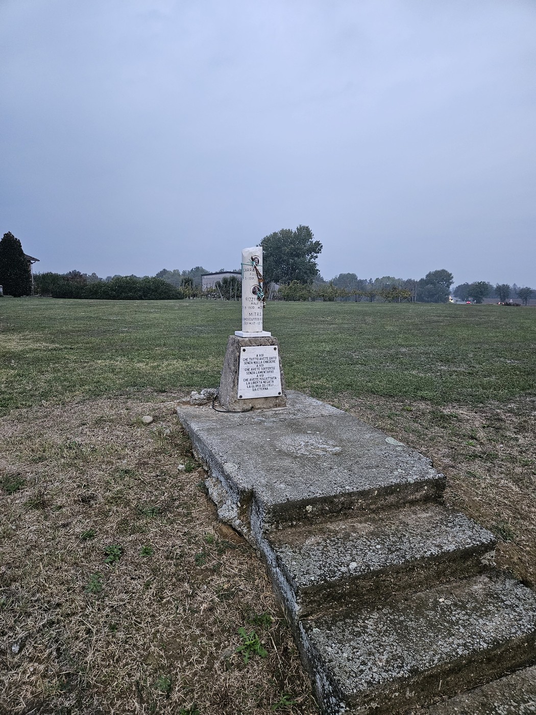 Cippo in pietra con iscrizione su base a gradini, isolato in un prato sotto cielo grigio.