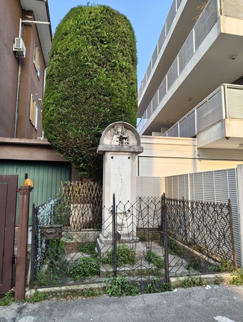 Il monumento ai caduti di Santa Bertilla, circondato da una recinzione, si staglia tra edifici moderni e vegetazione.