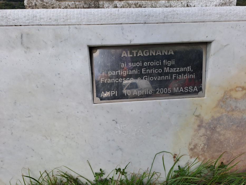 Targa metallica con iscrizione dedicata ai partigiani, fissata su basamento in marmo, tra l'erba.