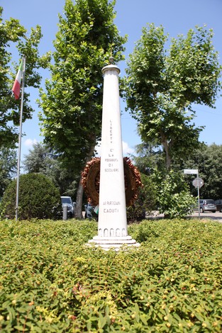 Monumento ai caduti circondato da alberi verdi, con una corona di fiori sulla base, in un contesto urbano.
