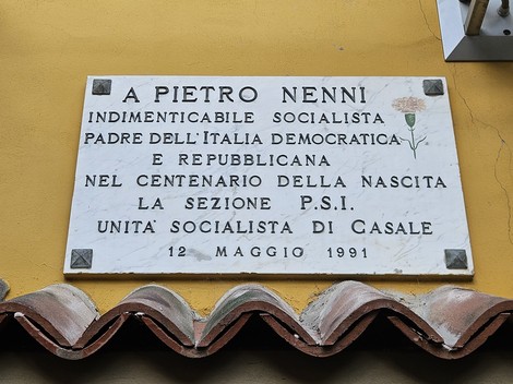 Lapide bianca con iscrizione dedicata a Pietro Nenni, fissata su una parete gialla sopra un tetto in coppi.