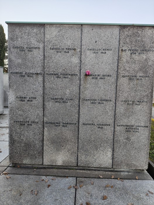 Monumento commemorativo con nomi di caduti, commemorato da un fiore rosa in un ambiente sereno.
