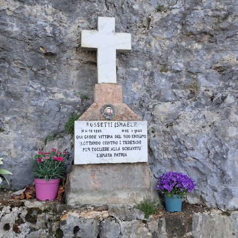 Cippo commemorativo a Rossetti, con iscrizioni e fiori colorati ai suoi piedi, situato su una roccia.