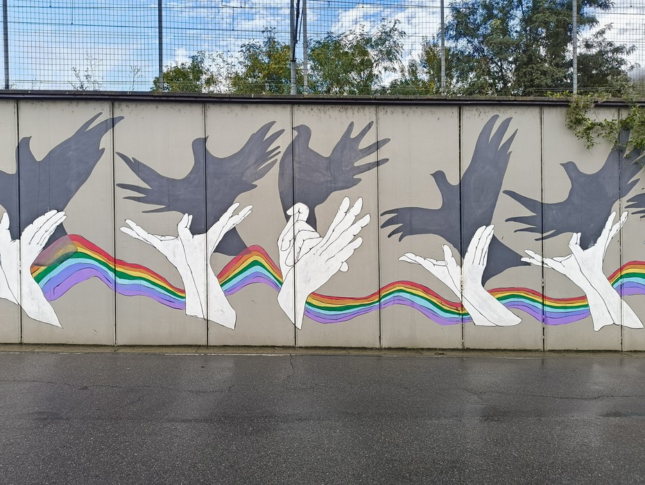 Murale di Pioppe con mani stilizzate e uccelli neri su sfondo grigio, arricchito da un arcobaleno vibrante.
